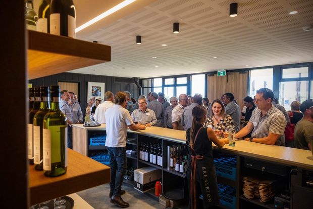A busy contemporary wine bar or hospitality venue with patrons gathered around a long timber-topped service counter. Built-in open shelving displays wine bottles and glassware, while dark cabinetry, timber wall panels, large windows, and a patterned suspended ceiling define the space. Warm linear lighting and black cylindrical downlights create an inviting social atmosphere.