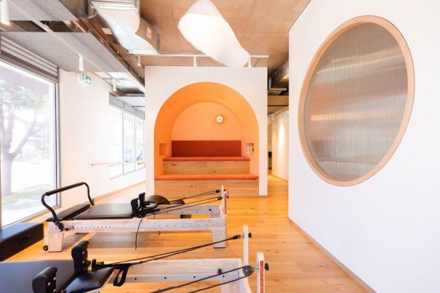 Bright contemporary Pilates studio with reformer equipment in the foreground, pale timber flooring, exposed services and ductwork, white walls, and a distinctive orange arched built-in alcove bench. A large oval ribbed-glass opening with a timber-toned frame forms a feature wall detail. The space has abundant daylight and an open, airy layout.