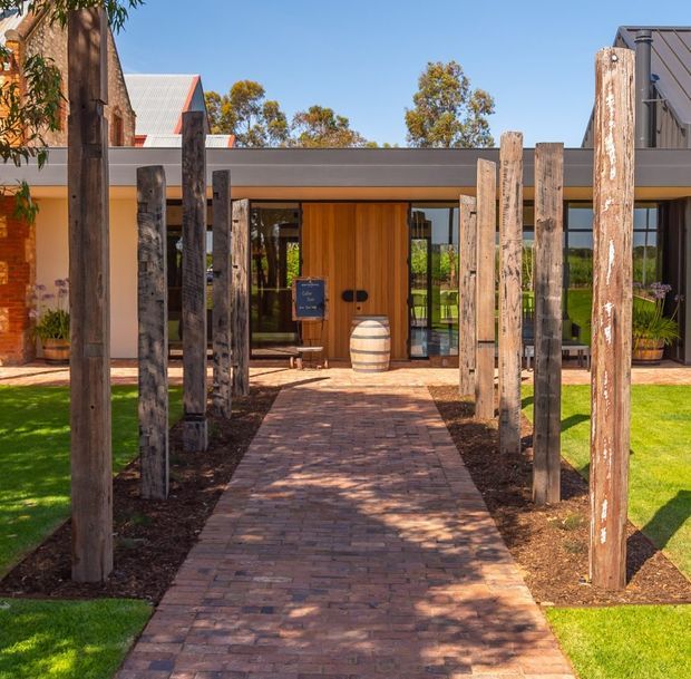 Exterior entrance to a low-rise contemporary building, approached by a brick-paved walkway framed with tall rough timber posts. The facade combines dark grey horizontal cladding, warm timber entry doors, extensive glazing, and sections of exposed red brick. Bright sunlight, manicured lawn, and native-style planting create a welcoming landscaped arrival.