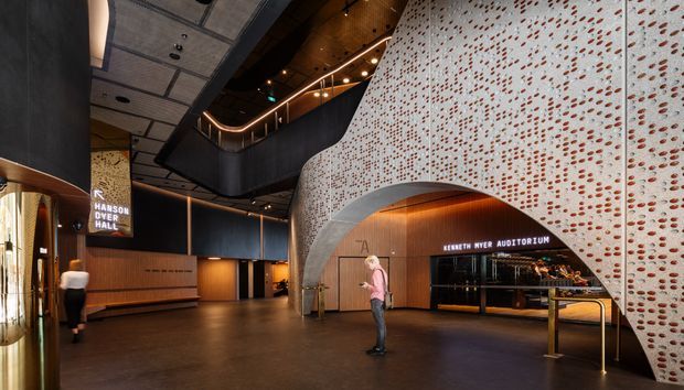 A dramatic contemporary performing arts venue foyer with a large curved perforated feature wall and arch framing the entrance to the Kenneth Myer Auditorium. The lobby includes warm timber wall panelling, dark metal surfaces, integrated linear lighting, a sculptural suspended ceiling, a reception or ticket counter, and directional signage to Hanson Dyer Hall.