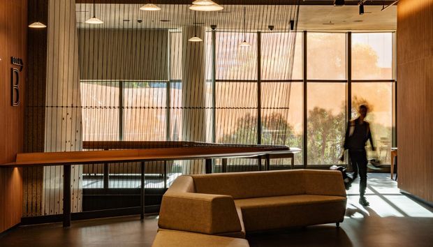 A warmly lit contemporary interior lounge or reception space with a long timber console or bar-height table, a low upholstered sofa, extensive floor-to-ceiling glazing, vertical timber screening, pendant lights, and dark exposed ceiling services. The space has a refined, inviting atmosphere with strong indoor-outdoor visual connection.