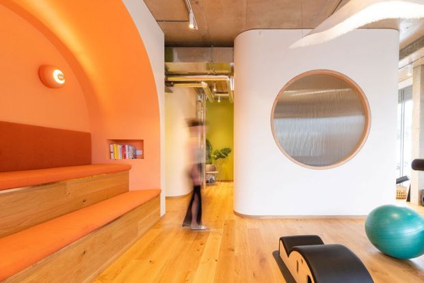 Vibrant contemporary interior with a sculptural white curved partition featuring a large round glazed opening. The space has an orange built-in stepped bench and arched alcove on the left, warm timber flooring, exposed concrete ceiling services, and a glimpse of a bright green room beyond. Modern lounge or communal workspace furnishings are visible in the foreground.