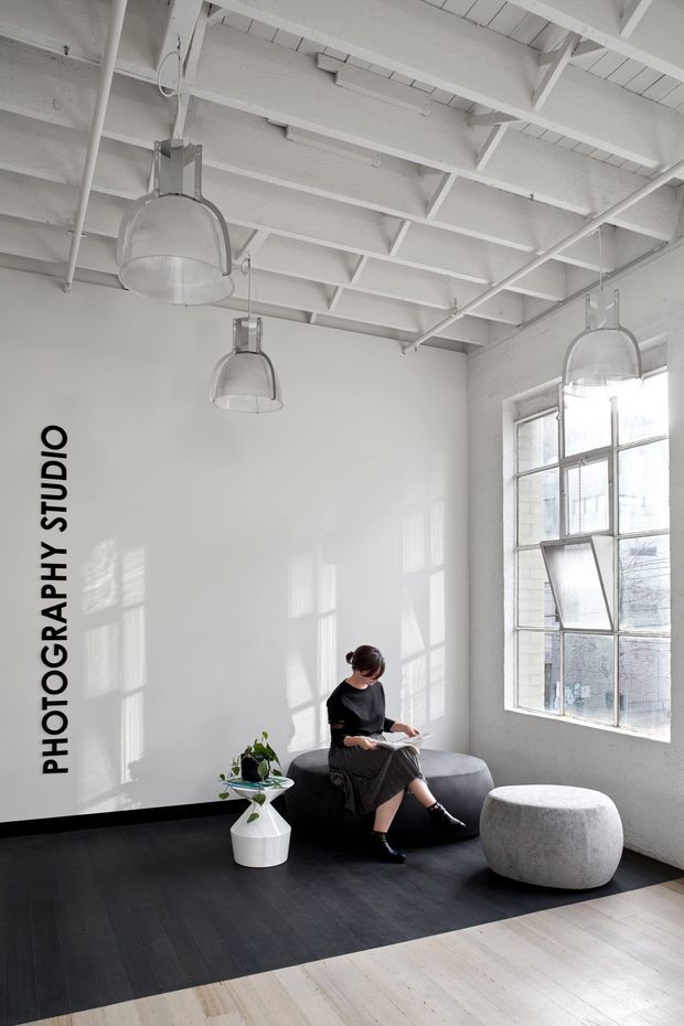 Bright, minimalist studio interior with white-painted exposed roof beams and rafters, large industrial-style multi-pane windows, and three suspended translucent pendant lights. A woman sits reading on a dark rounded sofa beside a small potted plant and a large rounded grey ottoman. The room has a dark timber floor area transitioning to pale timber flooring, with strong daylight and window shadows across the walls.