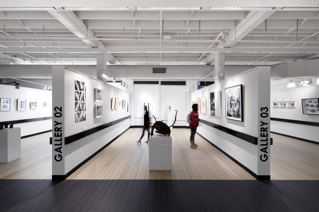 Bright contemporary art gallery interior with white walls, exposed white structural beams and services, light timber flooring, dark perimeter floor bands, framed artworks, and sculptural displays. The view looks through Gallery 02 toward Gallery 03, with visitors walking through the exhibition space.