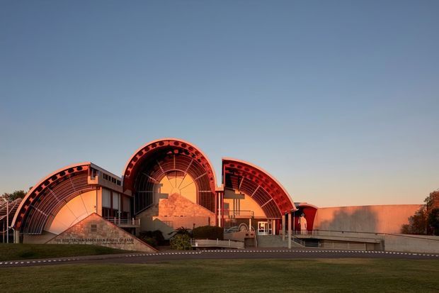 Exterior view of a monumental modern architectural complex at dusk, composed of several large curved barrel-vault structures with exposed ribbed framing and warm red-orange illumination. The building sits behind a broad lawn and paved forecourt, with a dramatic sculptural facade and clear sky.