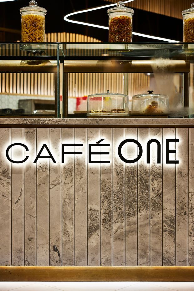 Close-up view of a contemporary café counter and display area. A backlit sign reading “CAFÉ ONE” is mounted on a vertically panelled, heavily veined stone or marble feature wall. Above the counter are glass display cases, shelves, jars of dried pasta or food products, and illuminated linear and pendant-style lighting. The palette is warm and refined, with cream stone, dark framing, brass-toned surfaces, and soft integrated lighting.