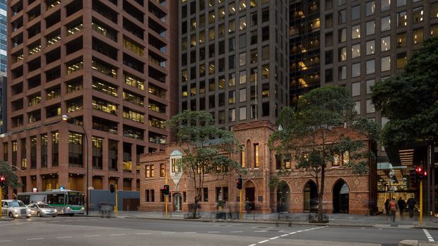 Street-level urban architectural photograph showing a historic red-brick Gothic Revival church or civic building retained between tall modern office towers. The scene includes a broad city roadway, traffic signals, buses, trees, and illuminated office windows at dusk.