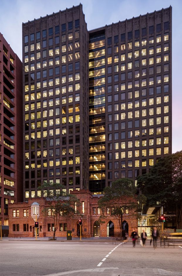Exterior architectural photograph of a tall urban commercial building at dusk, with a dark rectilinear masonry facade, repetitive grid of windows, and a glazed illuminated central atrium or vertical circulation zone. A lower historic red-brick building with arched openings and small tower-like details forms the street frontage, creating a contrast between heritage masonry and modern high-rise construction. The scene includes an urban roadway, traffic lights, street trees, and pedestrians.