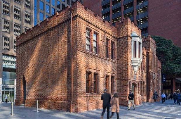 Urban exterior view of a historic red-brick masonry building with stepped parapets, tall narrow timber-framed windows, decorative stone window surrounds, and arched openings. The building sits within a dense modern city context, with contemporary high-rise buildings and pedestrians along the paved sidewalk.
