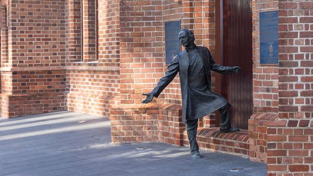 Exterior view of a red brick institutional or heritage-style building entrance, featuring a dark bronze statue of a person stepping through the doorway. The facade has projecting brick piers, recessed panels, plaques, and broad paved forecourt; no interior room is visible.