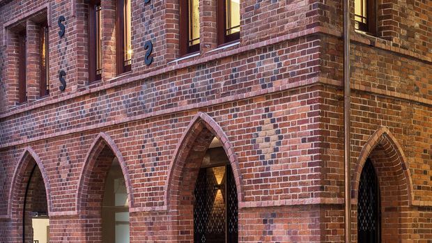 Close-up exterior view of a historic red-brick masonry building with pointed Gothic-arch openings, recessed dark metal doors or gates, narrow vertical windows, and decorative projecting brick courses. Warm architectural lighting highlights the textured brick facade and strong vertical and arched composition.