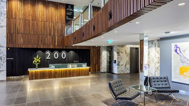 A spacious contemporary commercial lobby and reception area with a backlit timber reception desk, dark vertical timber feature wall, large illuminated “200” signage, marble-look wall cladding, polished tiled flooring, a mezzanine or upper-level balcony with timber slatted detailing, and a small leather lounge seating area around a glass coffee table. The space is bright, refined, and hotel-like.