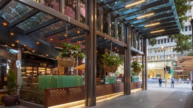 A sophisticated street-facing hospitality venue with a covered outdoor dining terrace and glazed pavilion structure. Dark steel framing supports a transparent canopy, with warm integrated lighting, hanging and potted greenery, patterned metal planter screens, and a turquoise tiled bar or counter visible inside. The urban commercial streetscape is visible beyond the terrace.
