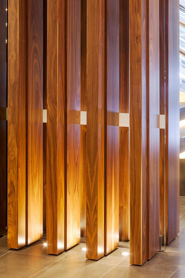 Interior architectural feature composed of tall, narrow vertical timber panels or screens with rich linear wood grain, arranged in parallel rows. Integrated warm uplighting illuminates the panels from the floor, while small light-coloured rectangular insert details punctuate the screen. The feature stands on large neutral stone or tile flooring, with glimpses of a contemporary interior and glazing at the right edge.