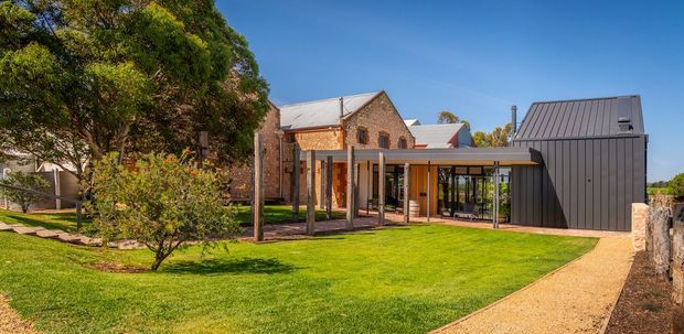 Exterior view of a renovated brick building with a contemporary dark metal-clad extension. The composition includes a pitched-roof brick structure, a long covered outdoor walkway or verandah with slender posts, extensive glazing, landscaped lawn, mature trees, and a gravel path. The design combines heritage-style masonry with a modern addition.