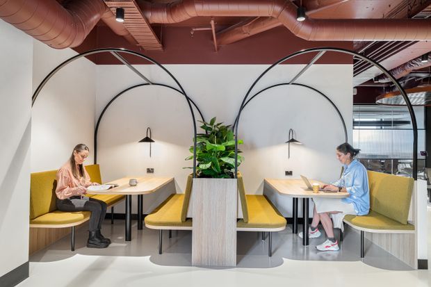 Contemporary commercial interior featuring symmetrical built-in booth seating in mustard yellow, light timber tables and planter joinery, black arched metal dividers, wall-mounted sconces, and exposed terracotta-painted ductwork and ceiling services. Two people are working at separate booths, with glazed meeting or office areas visible in the background.