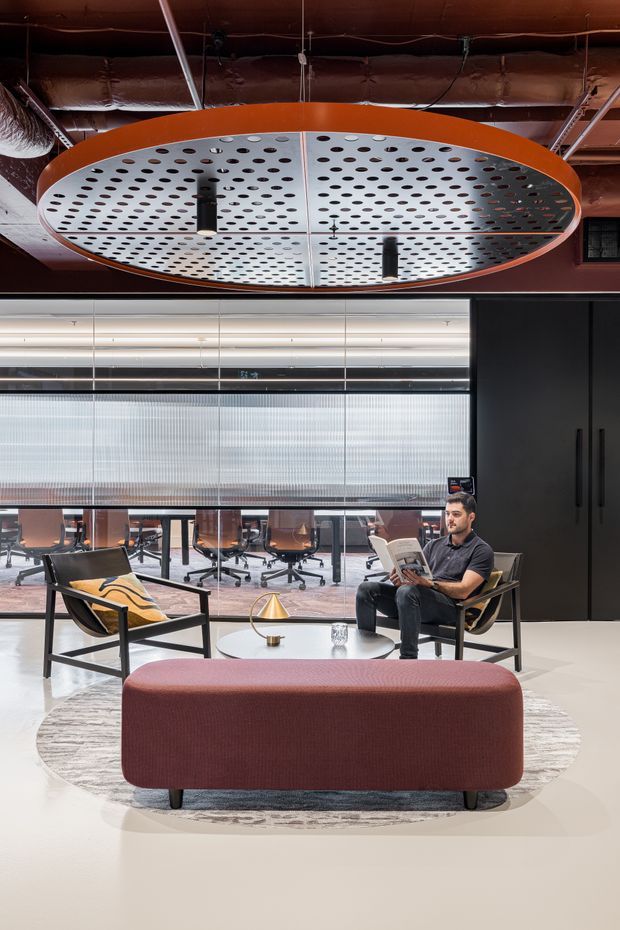 A contemporary commercial office lounge with a large circular perforated metal pendant light, exposed dark ceiling services, glass-partitioned workstations, and a relaxed seating area. The space includes black-framed lounge chairs, a burgundy upholstered ottoman, a round coffee table, and dark built-in cabinetry. The palette is modern and industrial with warm wood tones, polished light flooring, and muted burgundy accents.