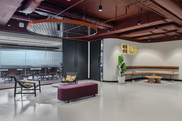 A contemporary commercial office reception and lounge area with an exposed services ceiling, polished light floor, curved feature wall, built-in timber bench seating, round tables, lounge furniture, and a visible meeting room behind glazed partitions. A branded wall sign and indoor planting complete the fit-out.