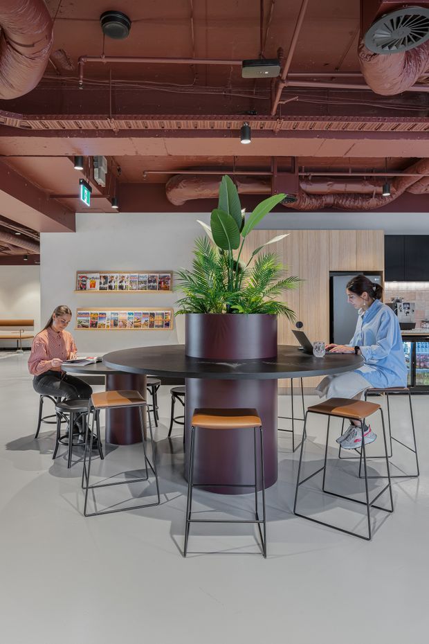 A bright contemporary commercial interior with a communal high table and integrated cylindrical planter surrounded by bar stools. Two people are working at the table, with magazine displays on the wall, a service or kitchenette area behind, and exposed painted ductwork, pipes, and ceiling services overhead. The space uses a restrained palette of light grey flooring, pale timber cabinetry, white walls, and dark burgundy-black furniture and metalwork.