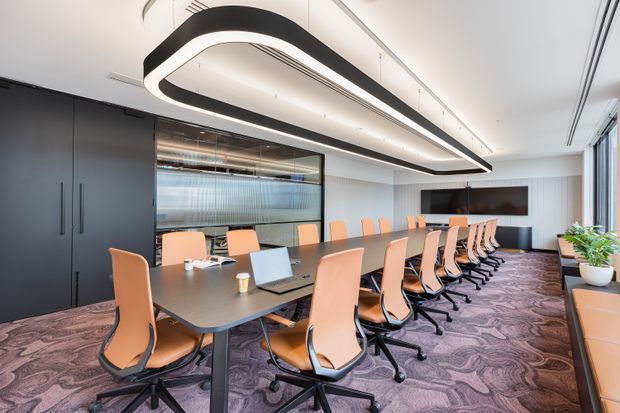 A contemporary commercial boardroom with a long dark conference table surrounded by orange upholstered swivel chairs. The room features dramatic linear sculptural ceiling lighting, dark built-in storage, a large wall-mounted display, glazed partitioning, full-height windows, and patterned carpet.