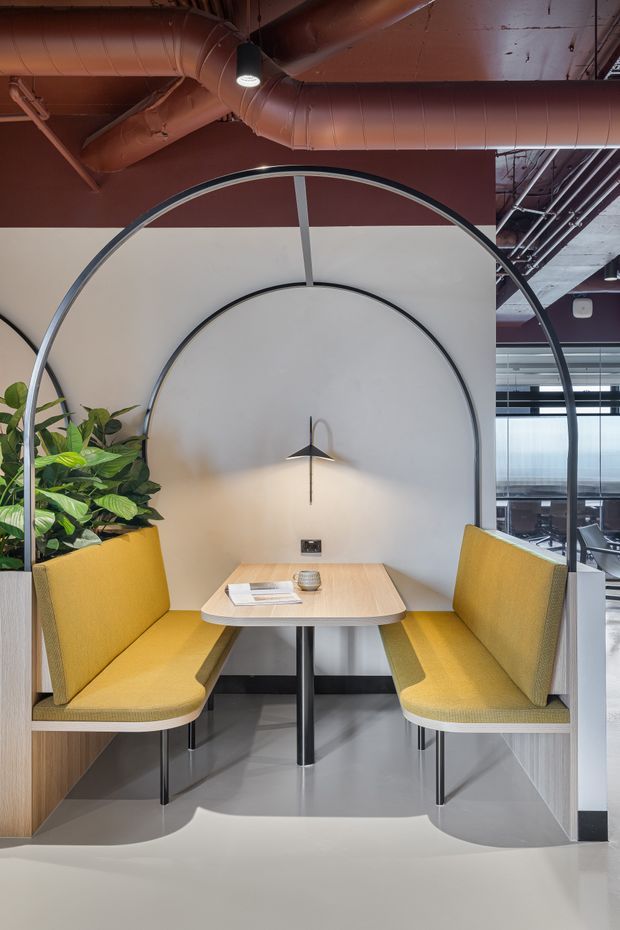 A contemporary commercial breakout or collaborative workspace featuring a compact booth with mustard-yellow upholstered bench seating on both sides of a light timber table. Large arched black metal frames create a distinctive canopy-like enclosure, with a sculptural wall light, indoor plants, polished light flooring, and exposed reddish-brown ceiling ductwork. Glazed office areas are visible in the background.