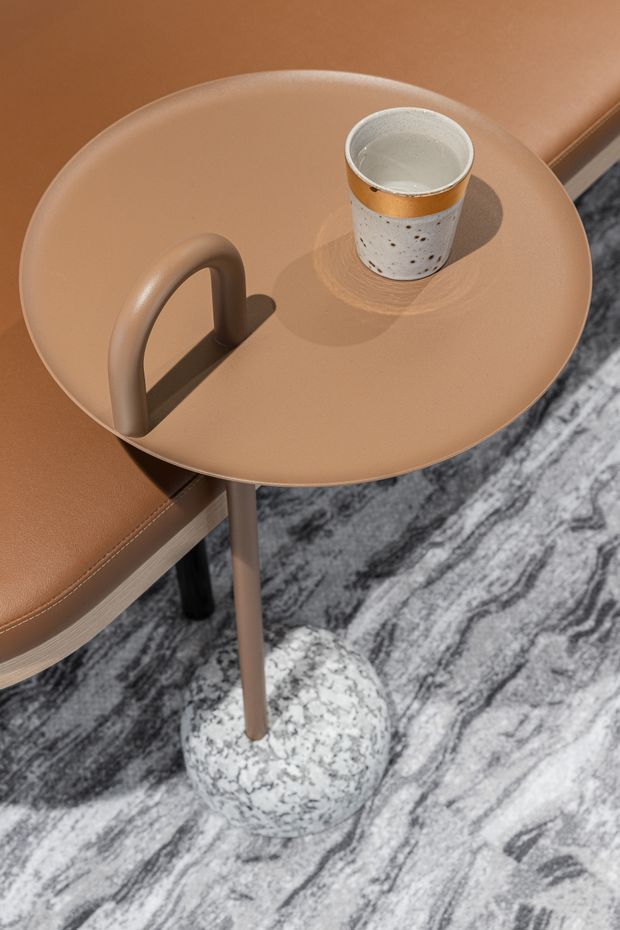 Close-up product-style image of a small round tan side table with an integrated loop handle, slim central leg, and mottled white stone-like spherical base. It sits beside a matching tan upholstered seat on a grey-and-white marbled rug, with a speckled ceramic cup placed on the tabletop.