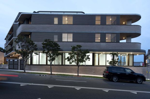 Street-level exterior view of a contemporary multi-storey building with rounded projecting balconies, dark horizontal louvred screening, extensive glazing at ground level, and a curved roofline detail. Small street trees and a parked vehicle sit in front of the facade.