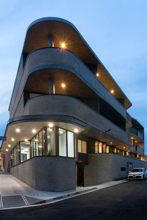 Street-level dusk view of a contemporary multi-storey residential or mixed-use building on a corner site. The building has strongly curved projecting balconies, dark grey masonry or concrete-like walls, extensive ground-floor glazing, warm recessed lighting, and a sweeping curved roof or canopy edge. A paved footpath and street are visible in the foreground.
