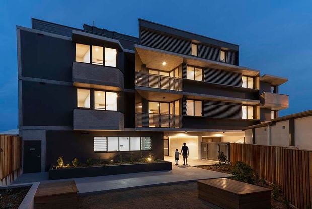 Twilight exterior view of a contemporary multi-storey residential apartment building. The dark rendered facade is articulated with projecting balconies, horizontal metal balustrades, timber-toned soffits, louvred screening, and warm illuminated glazing. A landscaped pedestrian entry court with timber fencing and built-in bench seating occupies the foreground.