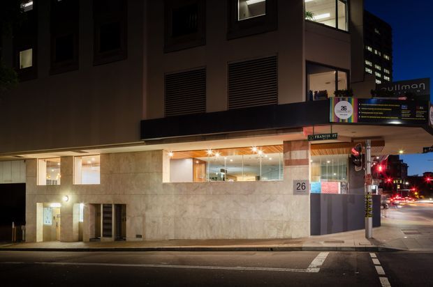 Night-time street view of a multi-storey commercial or hotel building at an urban intersection. The facade combines large pale stone-clad lower walls, dark upper walls, recessed windows, horizontal louvred ventilation panels, and illuminated ground-floor glazing. A covered entrance and projecting canopy sit beside a sign reading “Pullman”; street signage, traffic lights, and adjacent illuminated buildings reinforce the dense city setting.