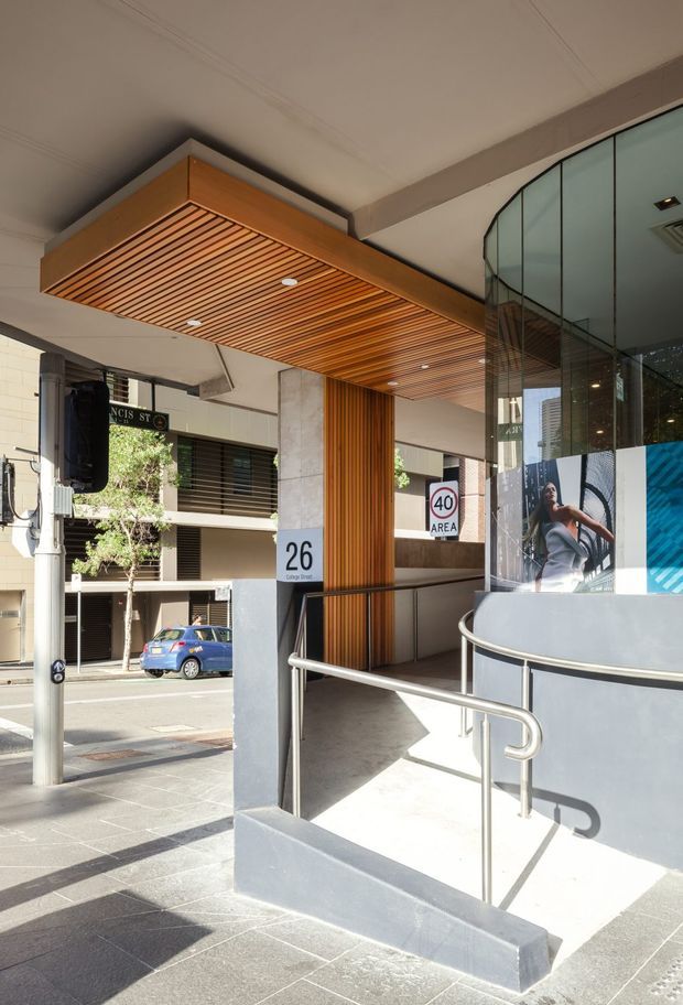 Street-level view of a contemporary commercial building entrance, featuring an accessible ramp with stainless-steel handrails, grey rendered walls, a large curved glass facade, and a prominent timber-lined soffit and vertical timber screen. Building number signage and urban streetscape are visible.