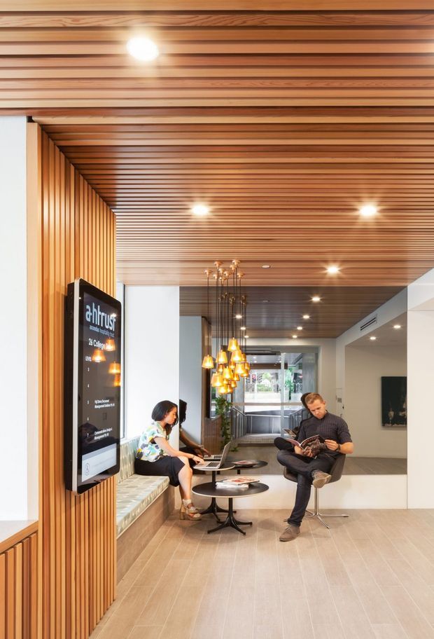 A bright contemporary commercial lobby or reception waiting area with a strong linear timber feature ceiling and matching vertical timber wall screening. The space includes a built-in bench beneath a large digital display, small round black tables, pendant lights, lounge seating, tiled flooring, and people using the waiting area. Glazing and an open circulation area are visible toward the rear.