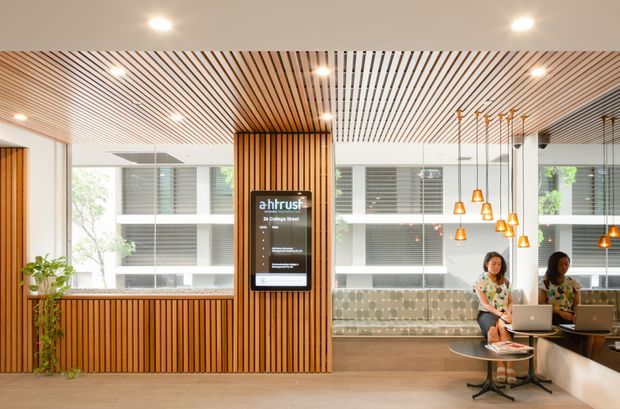 Bright contemporary commercial lounge or reception area with extensive vertical timber slat wall and ceiling detailing, integrated digital directory, large windows, patterned banquette seating, and pendant lights over small work tables. Two people are using laptops in the seating area.