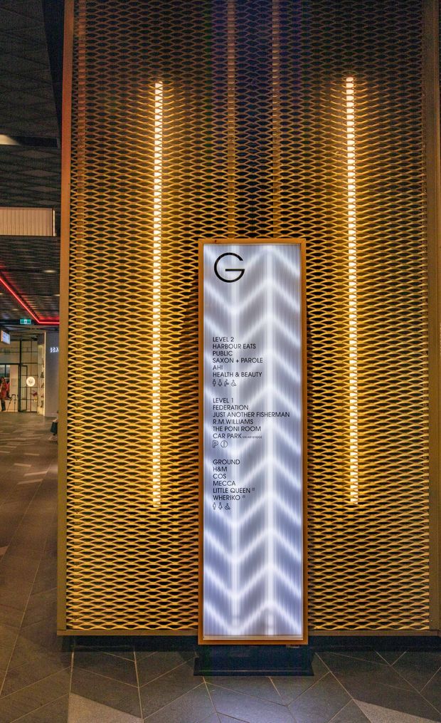 A dimly lit commercial interior lobby or shopping precinct featuring a tall gold-toned expanded-metal patterned feature screen, integrated vertical strip lighting, and a freestanding illuminated directory sign. Dark stone floor tiles and adjacent retail circulation areas are visible.