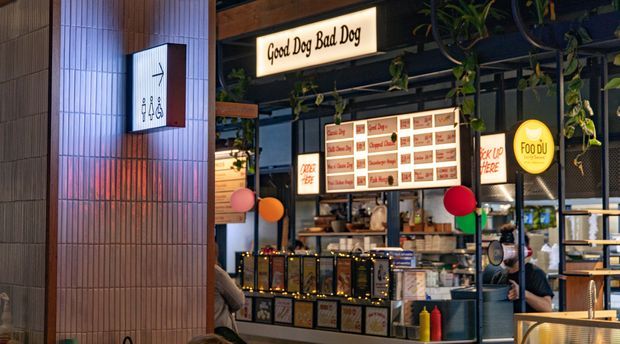 A warmly lit food-court or casual dining venue featuring a prominent Good Dog Bad Dog counter, illuminated menu boards, hanging greenery, a tiled column with directional signage, and visible seating and service counters. The scene has a contemporary, urban hospitality fit-out with colourful accent lighting and signage.