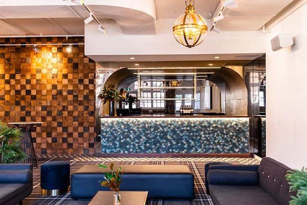 A stylish hospitality reception or lounge area with a patterned tiled reception counter, richly textured wood feature wall, arched metal-framed opening, plaid carpet, dark blue upholstered seating, indoor plants, and a prominent spherical brass pendant light. The space has a layered, eclectic contemporary character with warm ambient lighting.