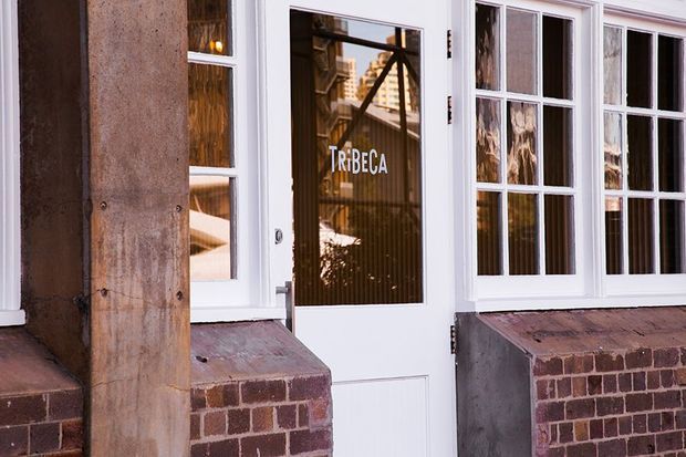 Close-up exterior view of a traditional storefront or building facade with a white glazed entry door and multi-pane windows set into a red-brown brick base. A weathered timber post stands beside the entrance, and the glazing reflects an urban streetscape. The word “TRIBECA” appears on the door.