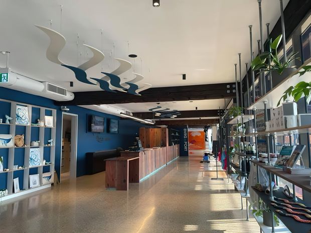 A bright, elongated commercial interior resembling a gallery, visitor centre, or retail reception space. The room has polished terrazzo-style flooring, deep blue walls, timber reception counters and cabinetry, open display shelving, extensive glazed frontage, and sculptural suspended ceiling elements in white and dark blue. Track and pendant lighting, shelving with displayed objects, and a long circulation aisle create a contemporary showroom-like environment.