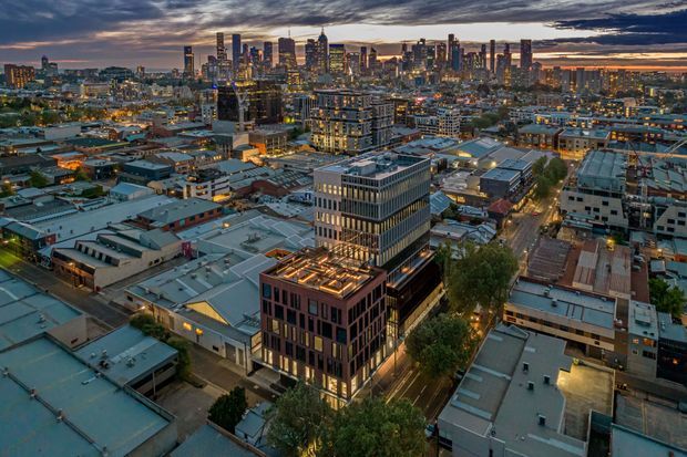 Aerial twilight view over a dense urban district, showing a mix of low-rise commercial and industrial buildings in the foreground and modern high-rise towers forming a city skyline in the distance. Streets, illuminated buildings, and varied rooflines create a layered metropolitan scene beneath dramatic cloud cover.