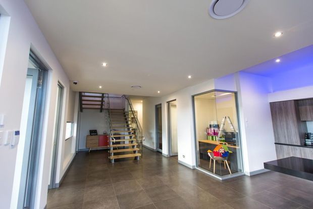 Open-plan contemporary interior with a tiled floor, white walls and recessed ceiling lighting. A modern open-riser timber and metal staircase leads to an upper level, while glazed partitions reveal a small office or work area. A kitchen with dark cabinetry and a black island is partially visible at right, accented by blue LED cove lighting.