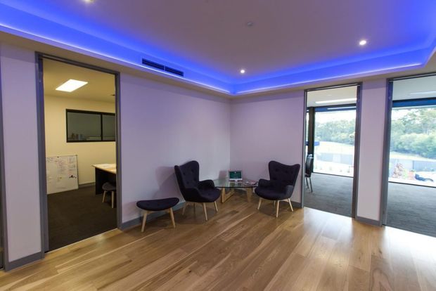 Contemporary office lounge or waiting area with timber flooring, two dark upholstered lounge chairs and matching ottomans, and blue LED cove lighting around the ceiling perimeter. Glazed openings connect to adjacent office spaces, with large windows providing views outside.