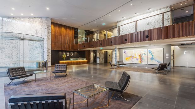 A spacious, double-height contemporary commercial lobby or hotel reception area with a polished grey tiled floor, dramatic veined marble feature wall, timber reception desk and wall panelling, mezzanine-level timber balustrade, glass partitions, modern lounge seating, and large abstract artworks. The space is refined and luxurious with strong architectural lighting and an open atrium-like volume.