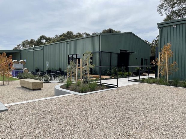 Exterior view of a contemporary low-rise building complex with dark green vertical metal cladding, simple pitched/gabled roof forms, black-framed glazing and an outdoor courtyard. The landscaped hardscape includes pale gravel, concrete paths, raised planting beds, young trees, a long concrete bench, outdoor tables and chairs, and black balustrades.