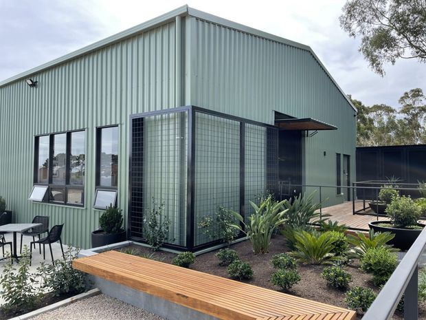 Exterior view of a contemporary single-storey building with pale green vertically ribbed metal cladding, a steep gabled roof, dark-framed windows, and a glazed screened entry or enclosed porch. A landscaped garden with raised planters and a timber bench occupies the foreground, with a paved outdoor seating area and deck visible beside the building.