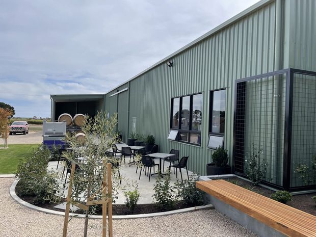 Exterior view of a contemporary green corrugated-metal building with a covered loading or storage bay, glazed entrance enclosure, and landscaped courtyard seating. The paved outdoor area includes café-style tables and chairs, raised timber seating, planter beds, and young olive trees. Industrial agricultural or commercial character with simple black-framed windows and doors.