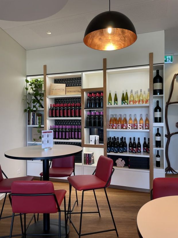 A contemporary wine-tasting retail and hospitality interior with white built-in display shelving filled with wine bottles, books, and boxed products. Burgundy upholstered chairs and small round tables sit on a warm timber floor. Vertical timber posts divide the shelving, while a large black pendant light with a copper-toned interior hangs overhead. The space is bright, polished, and inviting.