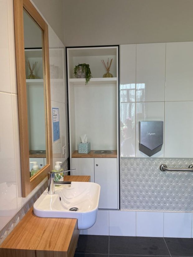 A bright, compact bathroom with a wall-mounted white basin, timber vanity countertop, large timber-framed mirror, white glossy wall tiles, a scalloped textured tile feature behind the basin, and integrated white cabinetry. A recessed open shelf displays small plants and reed diffusers, while chrome tapware and grab rails provide functional details. The dark tiled floor contrasts with the light walls and joinery.