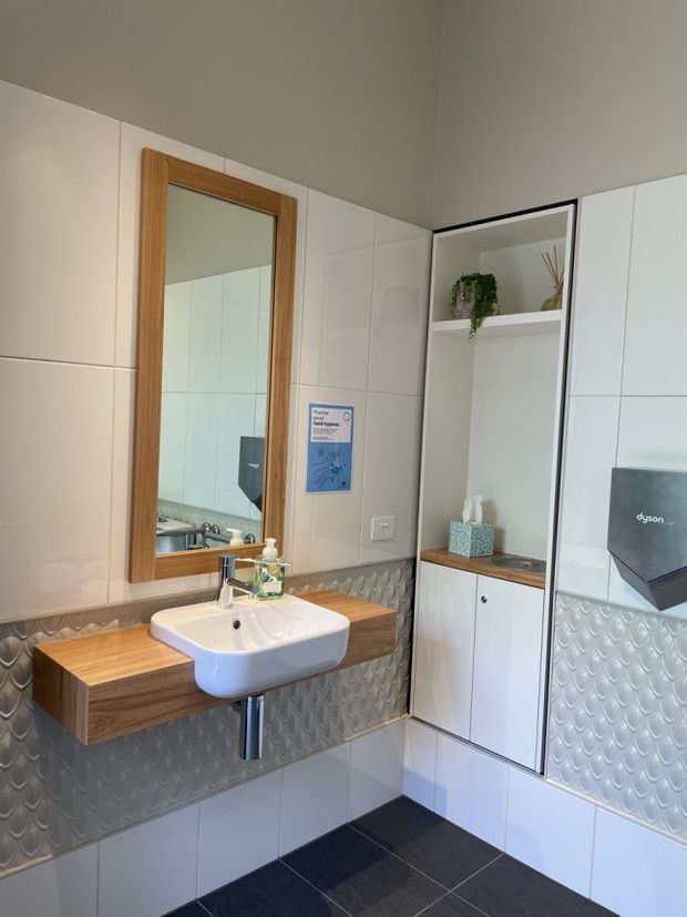 A compact contemporary bathroom with a wall-mounted white basin, timber floating vanity shelf, large timber-framed mirror, white wall tiles, and a grey patterned feature tile band. Built-in white cabinetry with open shelving and timber accents provides storage, while a wall-mounted Dyson hand dryer is visible on the right. The room has dark floor tiles and a clean, functional fit-out.