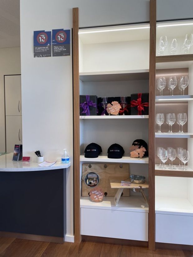 A contemporary retail or hospitality reception and display area. White built-in shelving framed with vertical timber members is illuminated by integrated strip lighting and displays wine glasses, gift boxes, caps, and branded promotional items. A dark reception counter with a pale stone-look top sits to the left, alongside white storage cabinetry. The space has warm timber flooring, clean white walls, and a bright, organised presentation.
