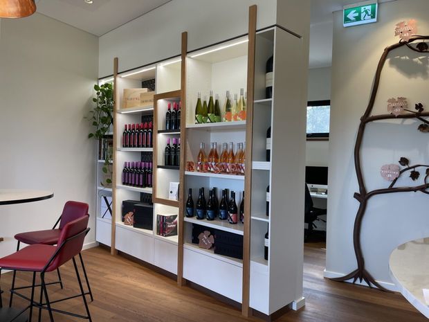 A modern retail or hospitality interior featuring a tall built-in display shelving unit stocked with bottles and products. The shelving is white with vertical timber accents and integrated lighting, set against warm timber flooring and pale walls. A small red seating area and round table sit in the foreground, while an adjacent room is visible through an opening. A decorative sculptural branch installation appears on the right wall, and an illuminated emergency exit sign is mounted near the ceiling.